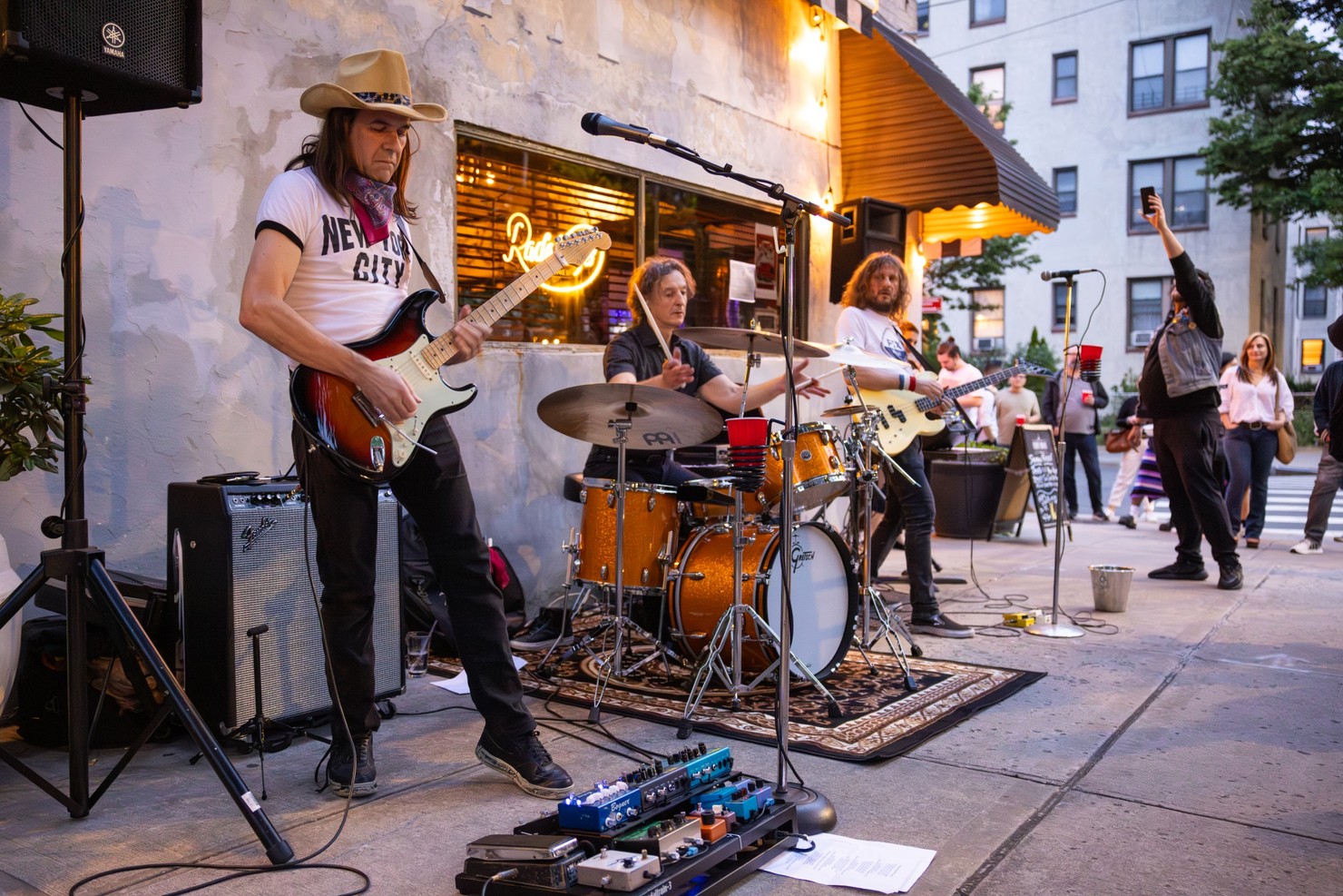 A guitarist, drummer, and bassist are playing their instruments outside a bar.