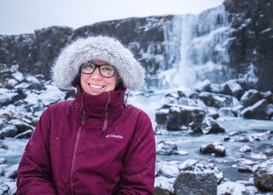 A person wearing a coat with a fuzzy hood standing in front of an icy waterfall.