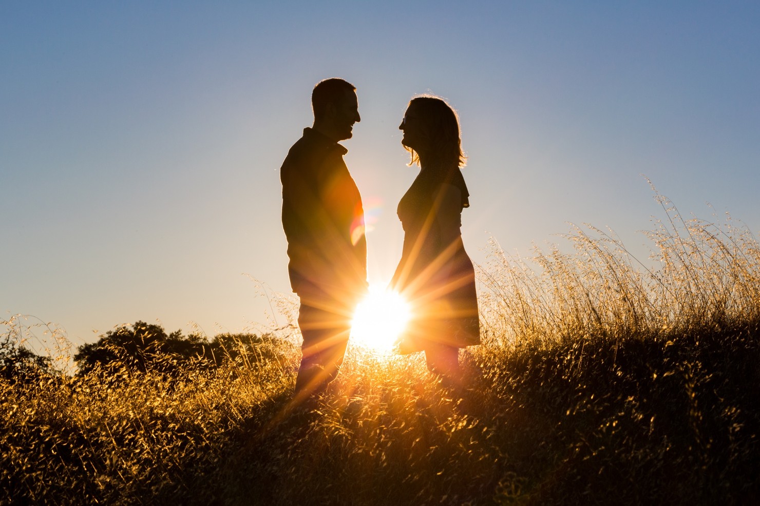 Two people standing close together with the sun setting between them.
