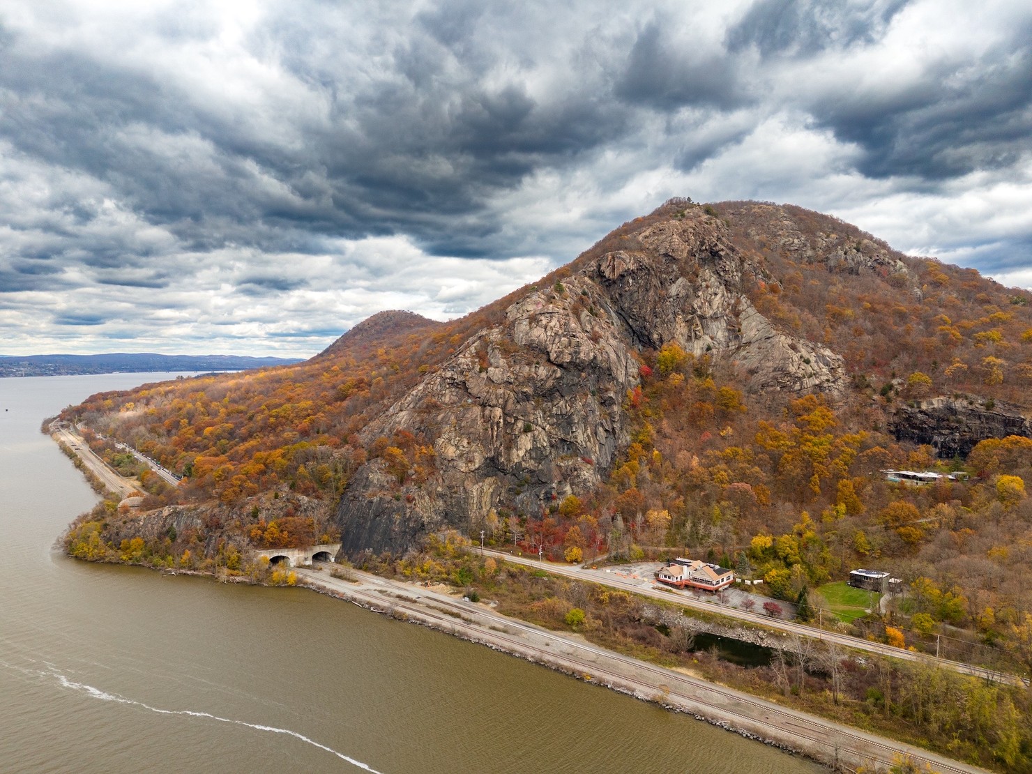 Breakneck Ridge as captured by drone, featuring a cloudy sky and orange leaves. Train tracks are in the foreground.