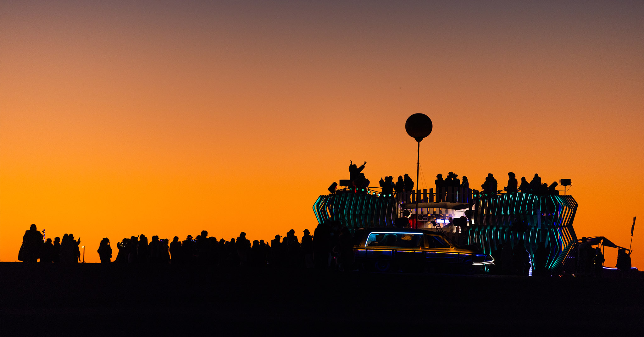 Several dozen people and a mobile music venue silhouetted against a sunset. The music venue is a large art car with a DJ booth on top.