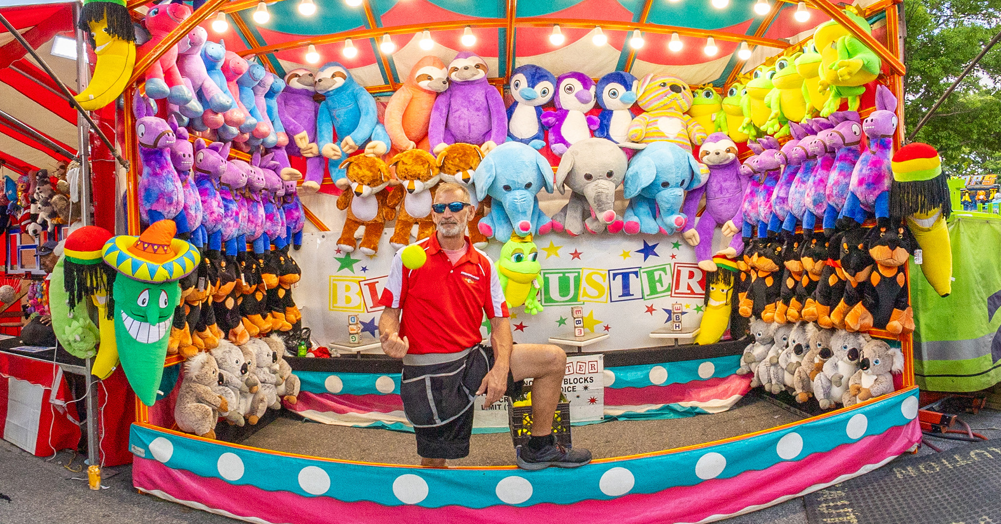 A wide angle photo of a man tossing a ball in the air in front of a carnival game.