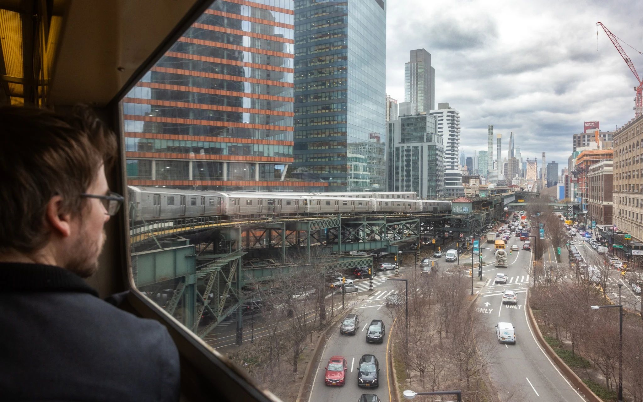 A man looking out a wide subway window at the Long Island City and Manhattan skylines.