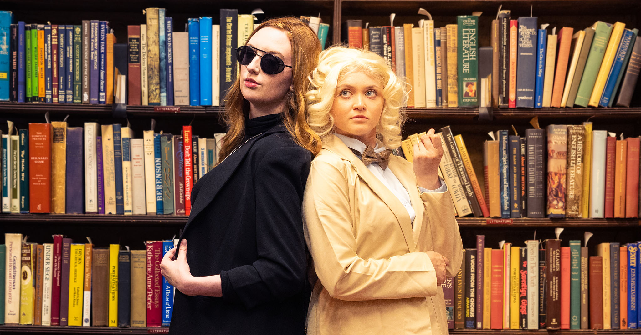 Good Omens' Aziraphale (Julia Duffy) and Crowley (Katie Gleason) standing back to back in front of a packed bookshelf.