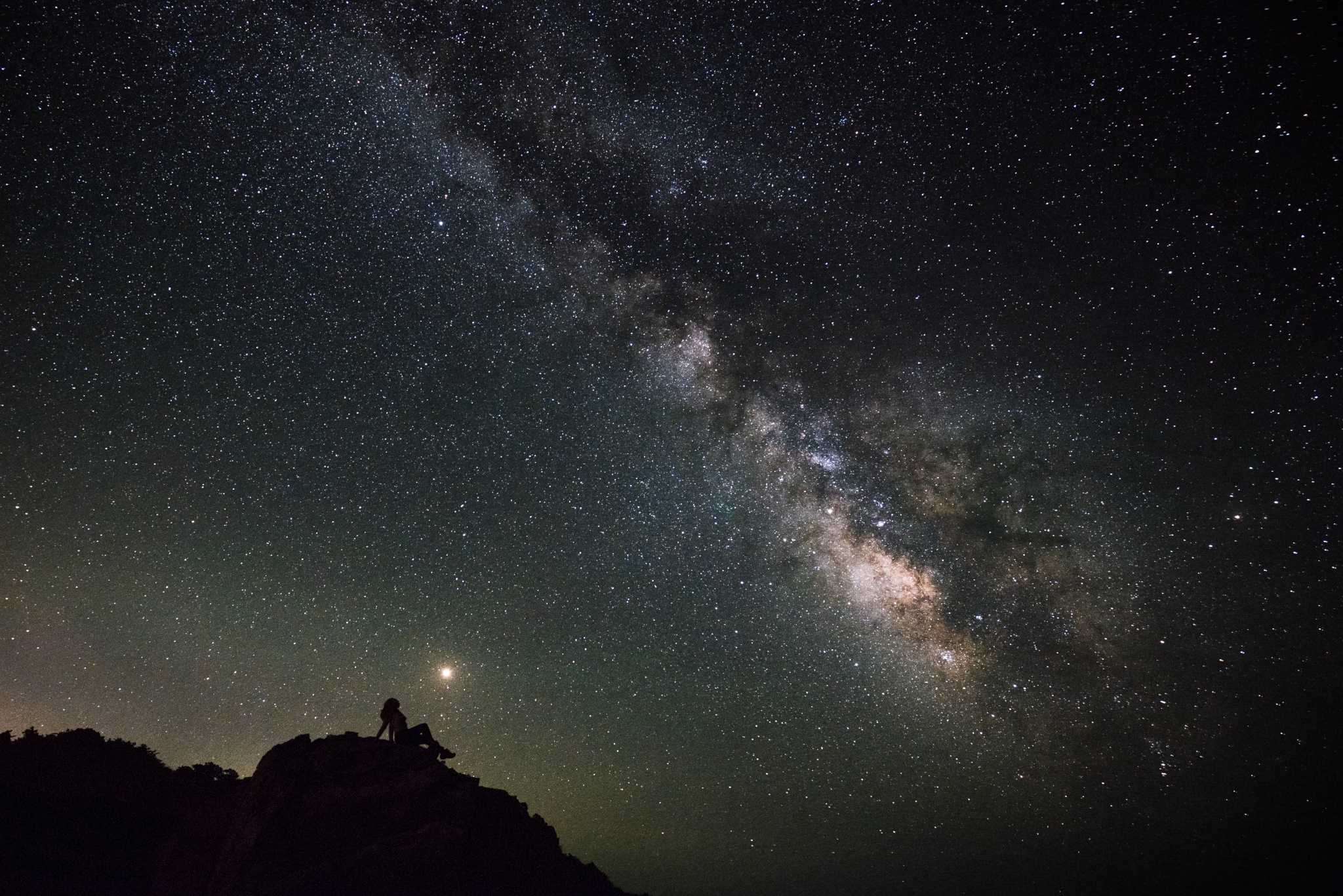 The Milky Way galaxy and Mars sit behind a person sitting on a rock in silhouette.